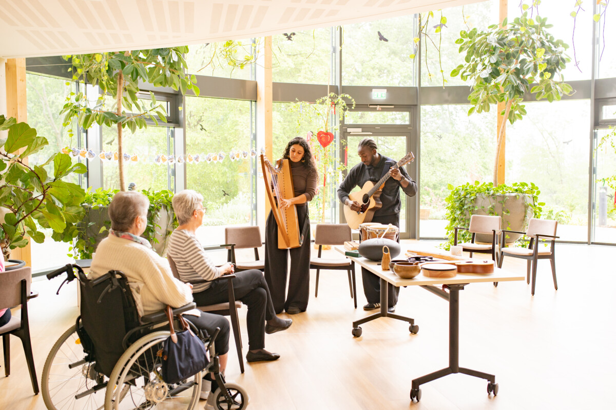 Zwei Musiker*innen und zwei älteren Frauen in einem hellen Raum mit großen Fenstern und vielen Pflanzen. Eine Person spielt Harfe, die andere Gitarre. Die älteren Frauen sitzen auf einem Stuhl und in einem Rollstuhl vor den Musizierenden. Auf einem Tisch weitere Musikinstrumente.