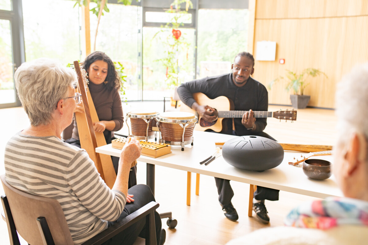 Zwei Musiker*innen und mehrere ältere Frauen sitzen in einem hellen Raum. Eine Person spielt Harfe, die andere Gitarre. Auf einem Tisch stehen und liegen verschiedene Musikinstrumente, darunter Trommeln, ein Xylophon, eine Steel Tongue Drum und Klangstäbe. Die älteren Frauen sitzen im Halbkreis vor den Musizierenden.