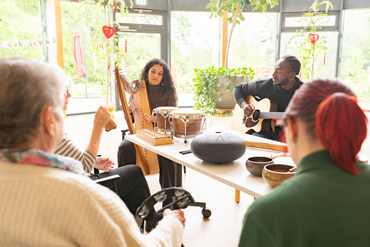 Foto von zwei Musiker*innen und mehreren älteren Frauen in einem hellen Raum mit großen Fenstern und vielen Pflanzen. Eine Person spielt Harfe, die andere Gitarre. Die älteren Frauen sitzen vor ihnen und halten Musikinstrumente in den Händen. Auf dem Tisch in der Mitte liegen weitere Instrumente.