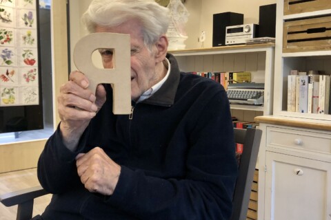 Ein älterer Mann hält lachend einen Pappbuchstaben vor sein Gesicht.