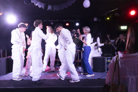 Mehrere Jugendliche in weißen Overalls bewegen sich tanzend auf einer Bühne in einem dunklen Raum mit bunten Scheinwerfern. Neben ihnen tanzt eine ältere Frau in Alltagskleidung. Im Hintergrund sind weitere Personen mit Mikrofon bzw. Filmkamera zu sehen.