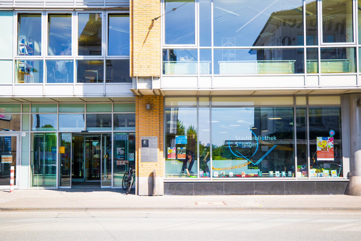 Außenansicht eines Gebäudes mit großen Glasfenstern und Eingangstüren. In der rechten Fensterfront steht: „Stadtbibliothek Solingen“. In der Mitte an der Wand hängt eine Tafel mit der Überschrift „EMIL KRONENBERG-HAUS“. Im Schaufenster liegen Bücher aus. Ein Fahrrad steht vor dem Eingang.