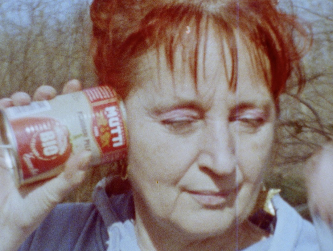 Selbstporträt der Künstlerin Dagie Brundert: Eine rotharige Frau hält sich eine geöffnete Dose mit rotem Etikett und der Aufschrift „BIO“ und „Tomaten“ ans Ohr. Im Hintergrund stehen kahle Bäume.