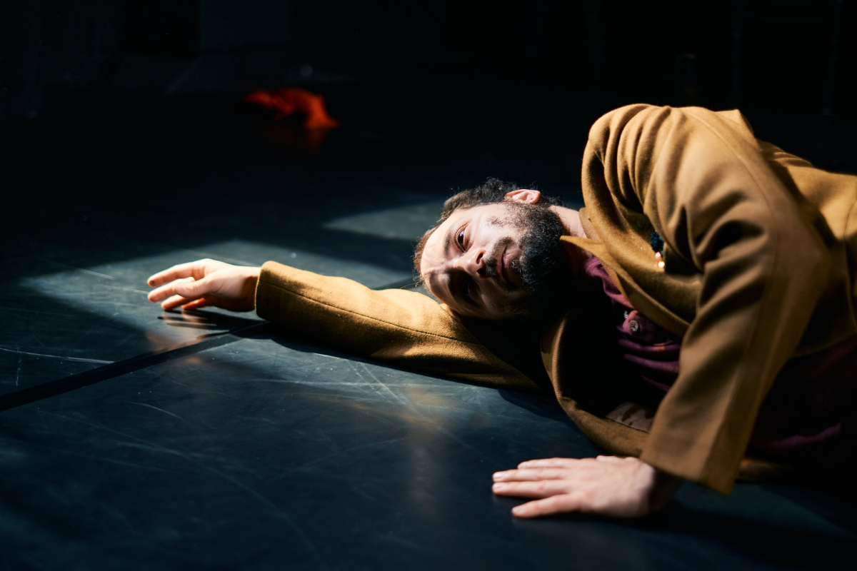 Ein Mann liegt seitlich auf einem dunklen Bühnenboden. Er trägt einen beigen Mantel. Sein Kopf liegt auf seinem ausgestreckten Arm.