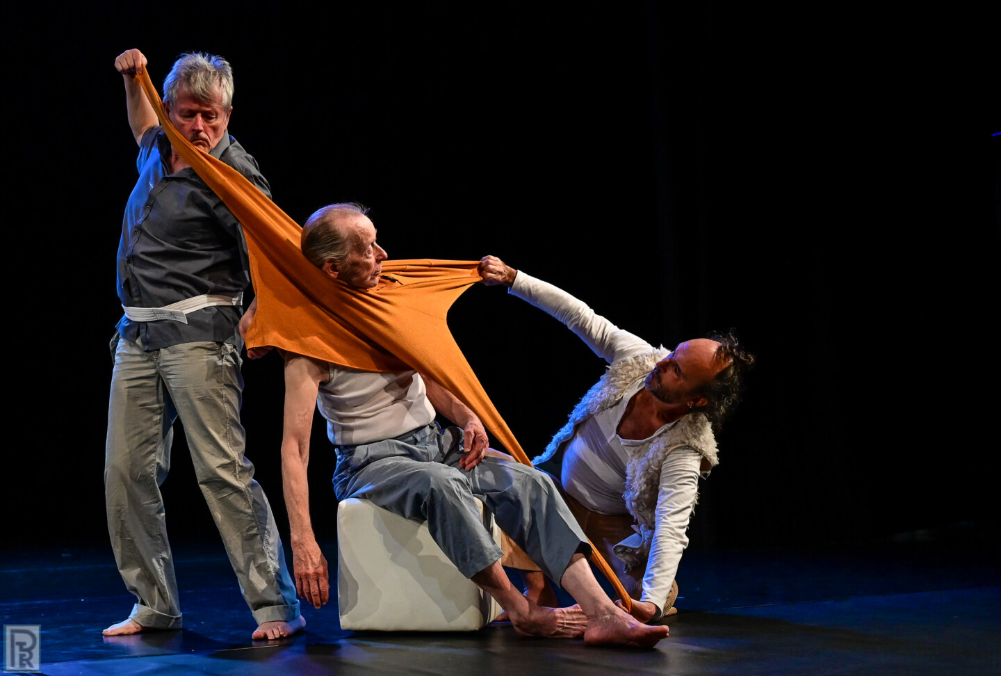 Drei Männer während einer Tanzperformance auf einer dunklen Bühne: Ein älterer Mann sitzt auf einem weißen Würfel. Er trägt eine orangefarbenes Oberteil um den Hals, ohne die Arme in den Ärmeln zu haben. Die beiden anderen Männer ziehen an dem Kleidungsstück, so dass der Stoff sich stark dehnt.