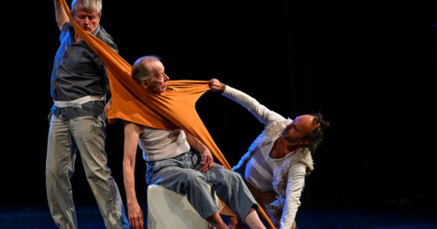 Drei Männer während einer Tanzperformance auf einer dunklen Bühne: Ein älterer Mann sitzt auf einem weißen Würfel. Er trägt eine orangefarbenes Oberteil um den Hals, ohne die Arme in den Ärmeln zu haben. Die beiden anderen Männer ziehen an dem Kleidungsstück, so dass der Stoff sich stark dehnt.