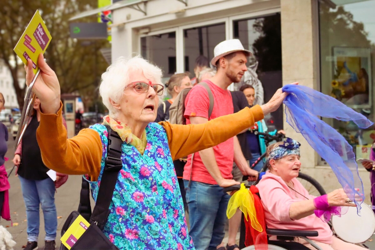 Eine ältere Frau in einer geblümten Kittelschürze steht mit ausgestreckten Armen auf der Straße und hält Flyer und ein violettes Tuch in den Händen. Daneben sitzt eine weitere ältere Person im Rollstuhl, der mit bunten Tüchern geschmückt ist. Sie trägt ein buntes Kopftuch hält ein Tamburin in der Hand.