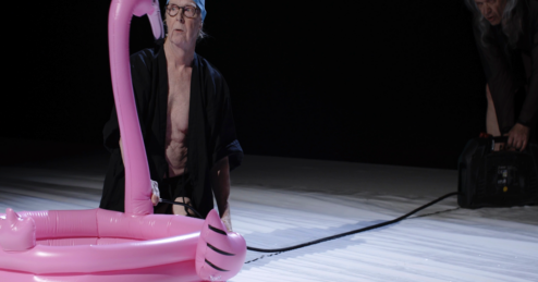 Eine ältere Person kniet auf einer hellen Bühne neben einem pinken Flamingo‑Planschbecken und trägt eine Badekappe und einen offenen Bademantel. Rechts bedient eine weitere ältere Person ein technisches Gerät.
