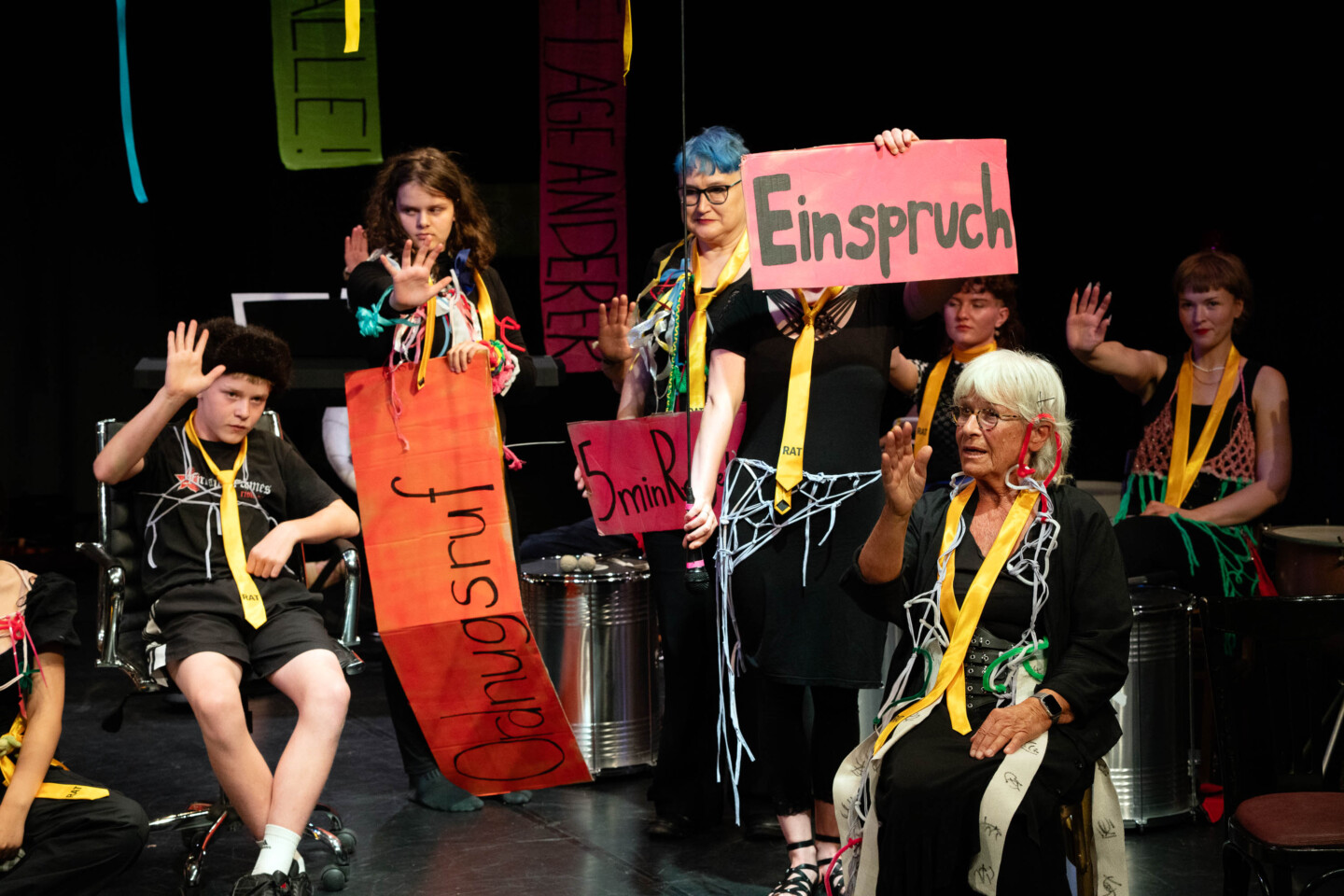 Auf einer Bühne sitzen und stehen mehrere junge und ältere Personen unterschiedlichen Alters. Sie tragen dunkle Kleidung, goldene Krawatten mit der Aufschrift „RAT“ und Teil bunte Bänder. Vorn sitzt eine ältere Frau mit weißem Haar. Mehrere Personen haben eine Hand erhoben. Einige halten Schilder mit Texten wie „Einspruch“ und „Ordnungsruf“.