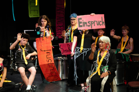 Auf einer Bühne sitzen und stehen mehrere junge und ältere Personen unterschiedlichen Alters. Sie tragen dunkle Kleidung, goldene Krawatten mit der Aufschrift „RAT“ und Teil bunte Bänder. Vorn sitzt eine ältere Frau mit weißem Haar. Mehrere Personen haben eine Hand erhoben. Einige halten Schilder mit Texten wie „Einspruch“ und „Ordnungsruf“.