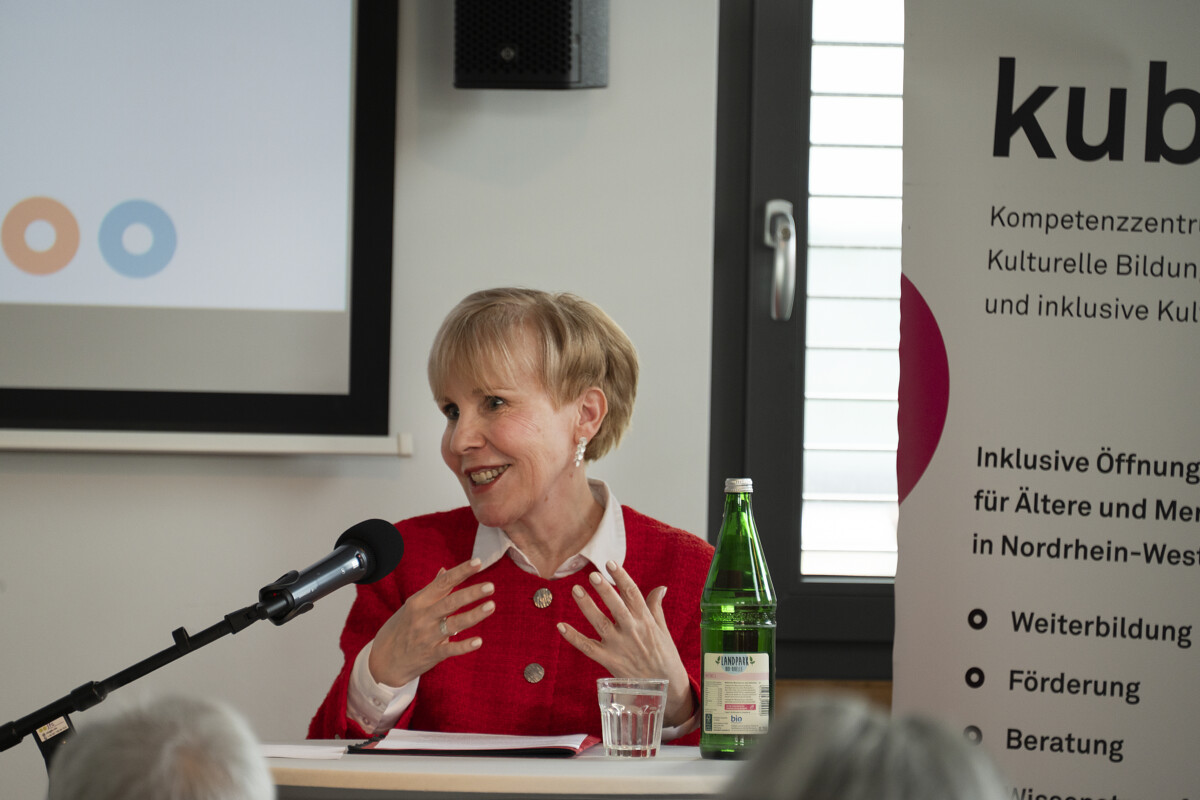 Claudia Determann, eine Frau mit kurzen blonden Haaren im roten Kostüm, steht hinter einem Stehtisch vor Publikum und spricht in ein Mikrofon. Auf dem Tisch stehen ein Glas und eine Wasserflasche.