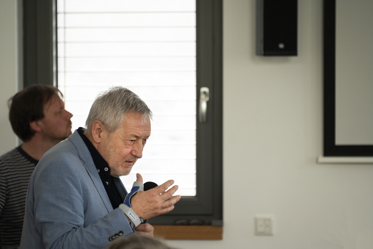 Frank Schulz-Nieswandt, ein Mann mit kurzem, grauem Haar in einem hellblauen Anzug, steht in einem seitlich zum Publikum, gestikuliert und spricht in ein Handmikrofon.