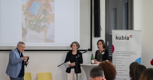 Ein Mann, Frank Schulz-Nieswandt, und zwei Frauen, Gundula Iblher und Barbara Scharfenberg, stehen unterhalb einer Projektionsfläche in einem Saal vor Publikum.