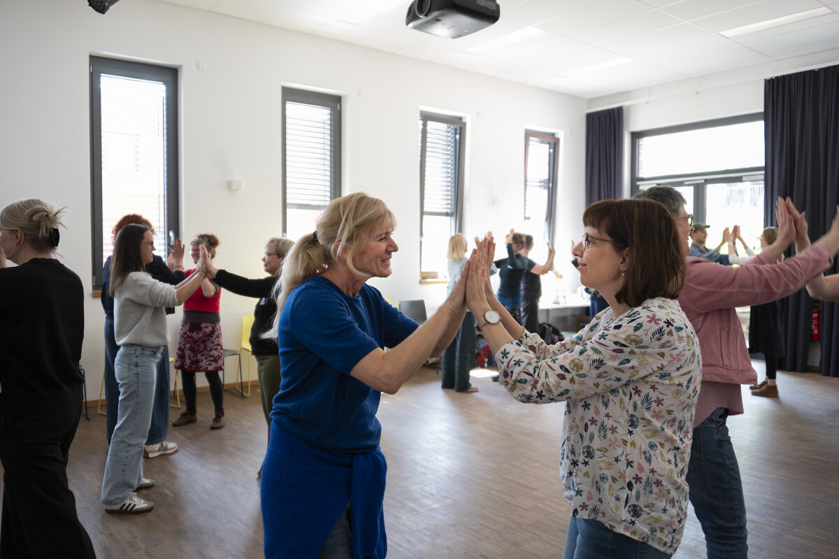 Zwei Frauen stehen sich gegenüber und berühren sich mit den erhobenen Handflächen. Im Hintergrund machen weitere Paare von Teilnehmende dieselbe Übung.