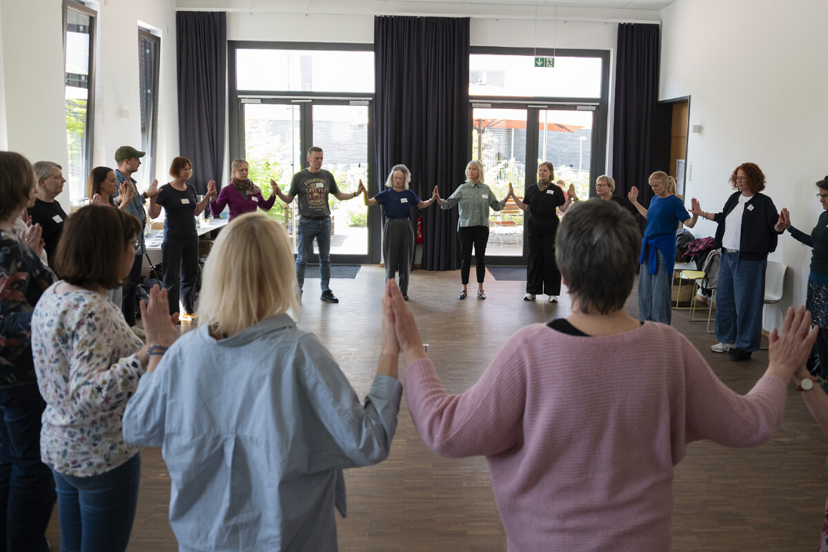 Eine größere Gruppe steht im Kreis in einem großen Raum. Die Teilnehmenden haben beide Hände jeweils auf Schulterhöhe angehoben, die Handflächen zeigen nach außen und liegen an den Handflächen der benachbarten Personen an. bzw. zu den jeweils benachbarten Personen. halten Abstand zueinander. Alle haben die Arme seitlich angehoben. Der Raum ist hell und leer geräumt.