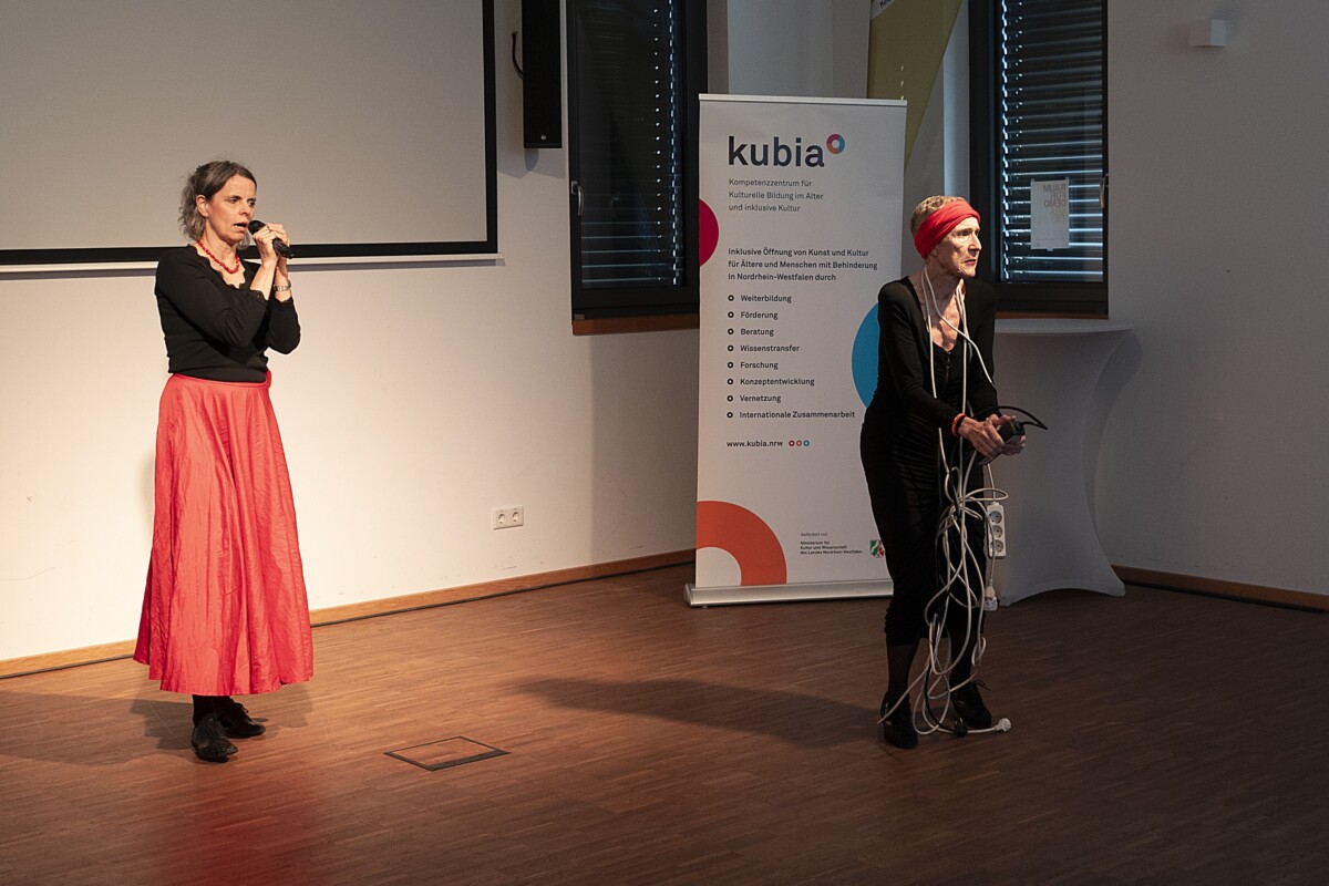 Zwei Frauen führen eine Performance auf. Die eine trägt einen langen roten Rock spricht in ein Mikrofons. Die andere trägt dunkle enge Kleidung und hält mehrere Verlängerungskabel i den Händen. Im Hintergrund steht ein kubia-Roll-up.