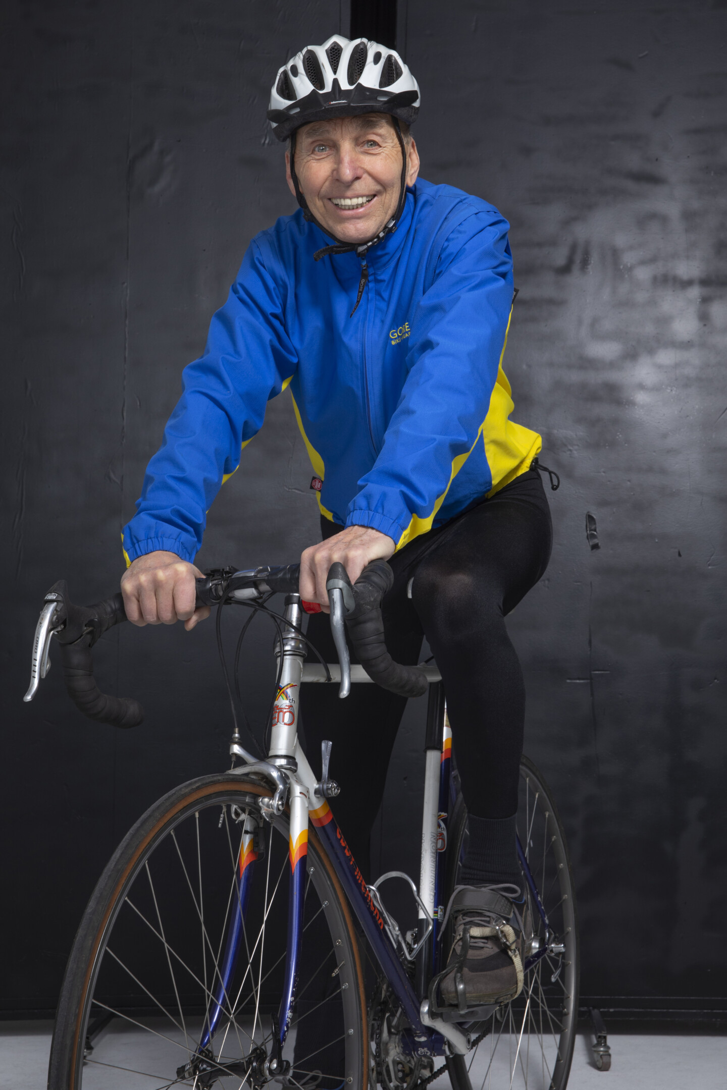 Ein älterer Mann sitzt auf einem Rennrad mit den Händen am Lenker und blickt lächelnd in die Kamera. Er trägt einen Fahrradhelm, eine blaue Jacke mit gelben Einsätzen und eine lange schwarze Radlerhose.