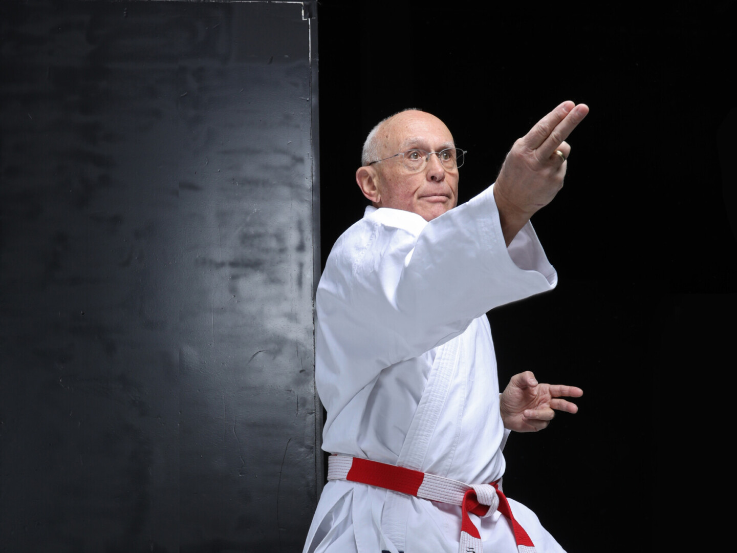 Eine älterer Mann steht barfuß in einer Karate-Pose mit gebeugten Knien und ausgestreckten Armen. Er trägt einen weißen Karateanzug mit rot-weißem Gürtel.