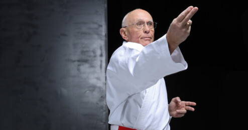 Eine älterer Mann steht barfuß in einer Karate-Pose mit gebeugten Knien und ausgestreckten Armen. Er trägt einen weißen Karateanzug mit rot-weißem Gürtel.