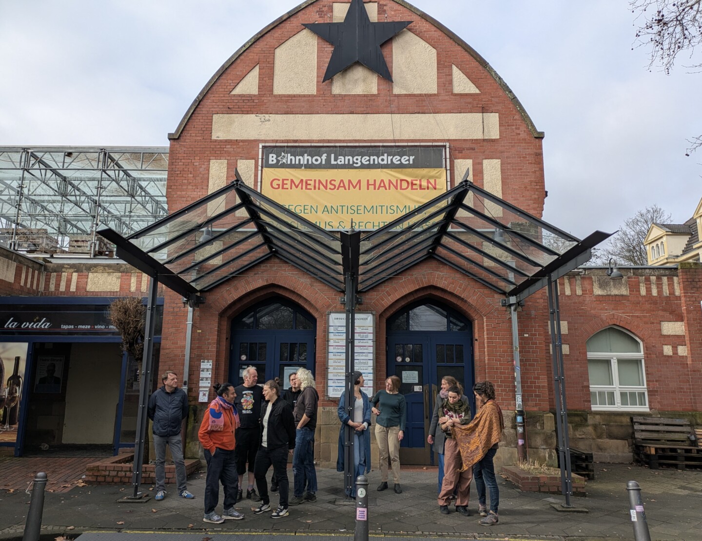 Foto von mehreren Personen, die vor dem Eingang eines Backsteingebäudes mit hohem Giebel und gläsernen Vordach stehen. Am Giebel hängt ein großer schwarzer Stern und darunter ein Banner mit der Aufschrift: „Bahnhof Langendreer GEMEINSAM HANDELN GEGEN ANTISEMITISMUS, RASSISMUS & RECHTEN HASS“.