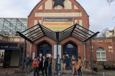 Foto von mehreren Personen, die vor dem Eingang eines Backsteingebäudes mit hohem Giebel und gläsernen Vordach stehen. Am Giebel hängt ein großer schwarzer Stern und darunter ein Banner mit der Aufschrift: „Bahnhof Langendreer GEMEINSAM HANDELN GEGEN ANTISEMITISMUS, RASSISMUS & RECHTEN HASS“.