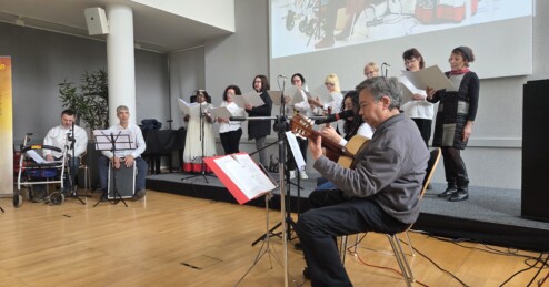 Das interkulturelle Ensemble von Wortklang bei einem Auftritt: Foto: Eine Gruppe musiziert gemeinsam. Im Vordergrund sitzt eine Person mit Saiteninstrument, im Hintergrund stehen Sänger*innen.