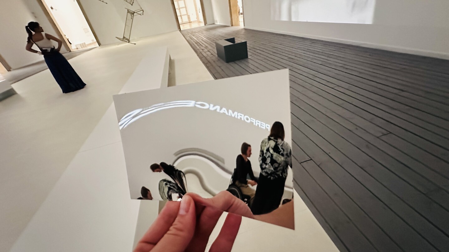 oto eines großen, nahezu leeren Ausstellungsraums mit weißen Wänden und einem zweigeteilten Boden: links ein heller, weißer Bereich, rechts dunkle Holzdielen. Im Vordergrund hält eine Hand eine quadratische Spiegel- oder Reflexionsfläche in die Kamera. Darin sind verzerrte Spiegelungen mehrerer Personen sowie geschwungene helle Linien zu sehen. Am oberen Rand der Spiegelung erscheint der spiegelverkehrte Schriftzug „PERFORMANCE“. Links im Raum steht eine Person in weiter dunkler Kleidung nahe einer Türöffnung.