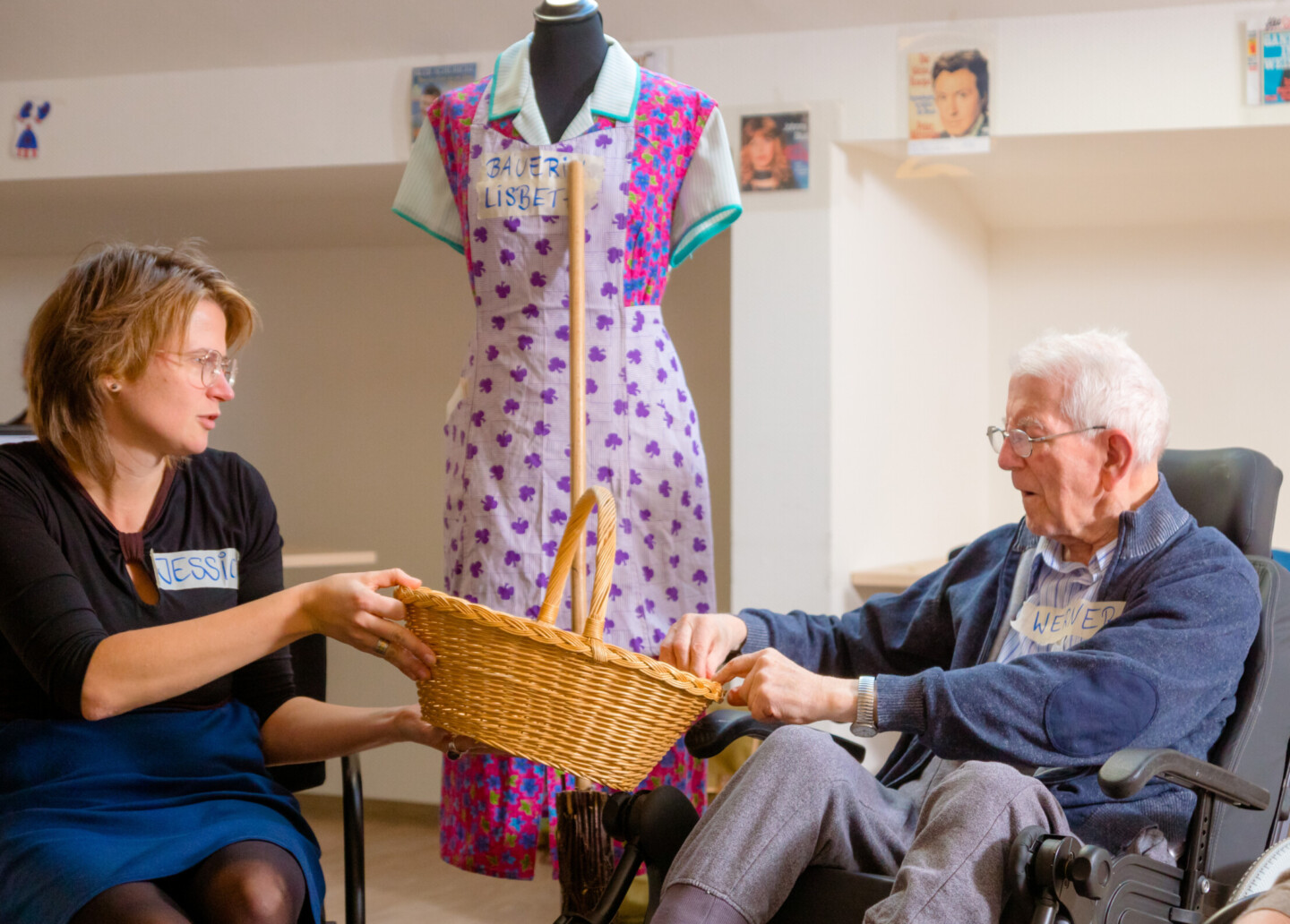Eine jüngere Frau, Jessica Höhn, und ein älterer Mann im Rollstuhl reichen sich einen geflochtenen Korb. Im Hintergrund steht eine Schaufensterpuppe in gemustertem Kleid und Kittelschürze.