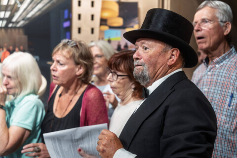 Rechts vorne steht ein bärtiger älterer Mann mit schwarzem Zylinder und schwarzem Sakko. Im Hintergrund sind ältere Menschen zu sehen, die sprechen oder singen